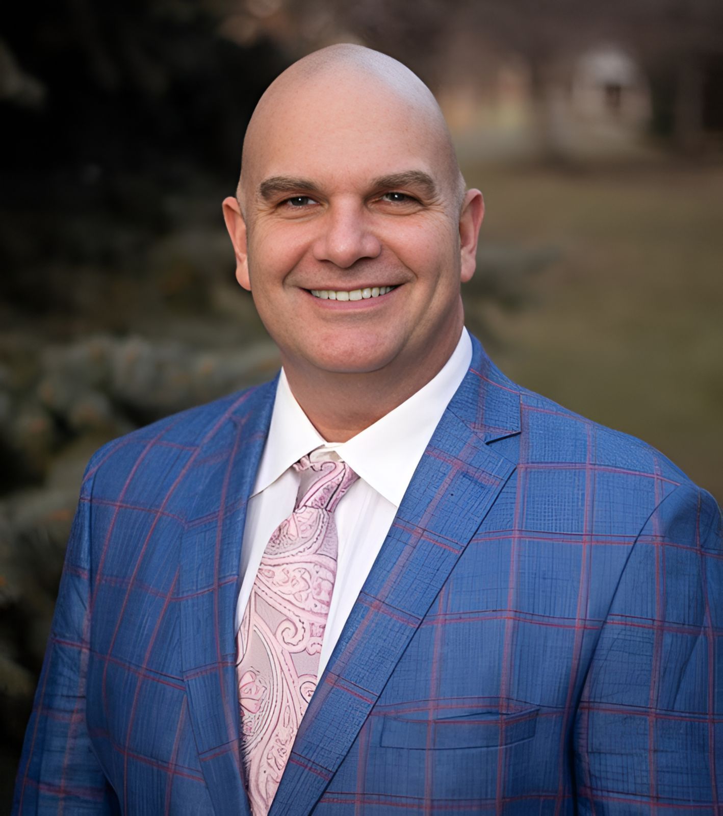 Man wearing a blue checkered suit with a pink tie against a blurred natural background